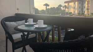 Un balcone con sedie in vimini, un tavolo di vetro, due tazze bianche e vista su un edificio con tetto rosso.