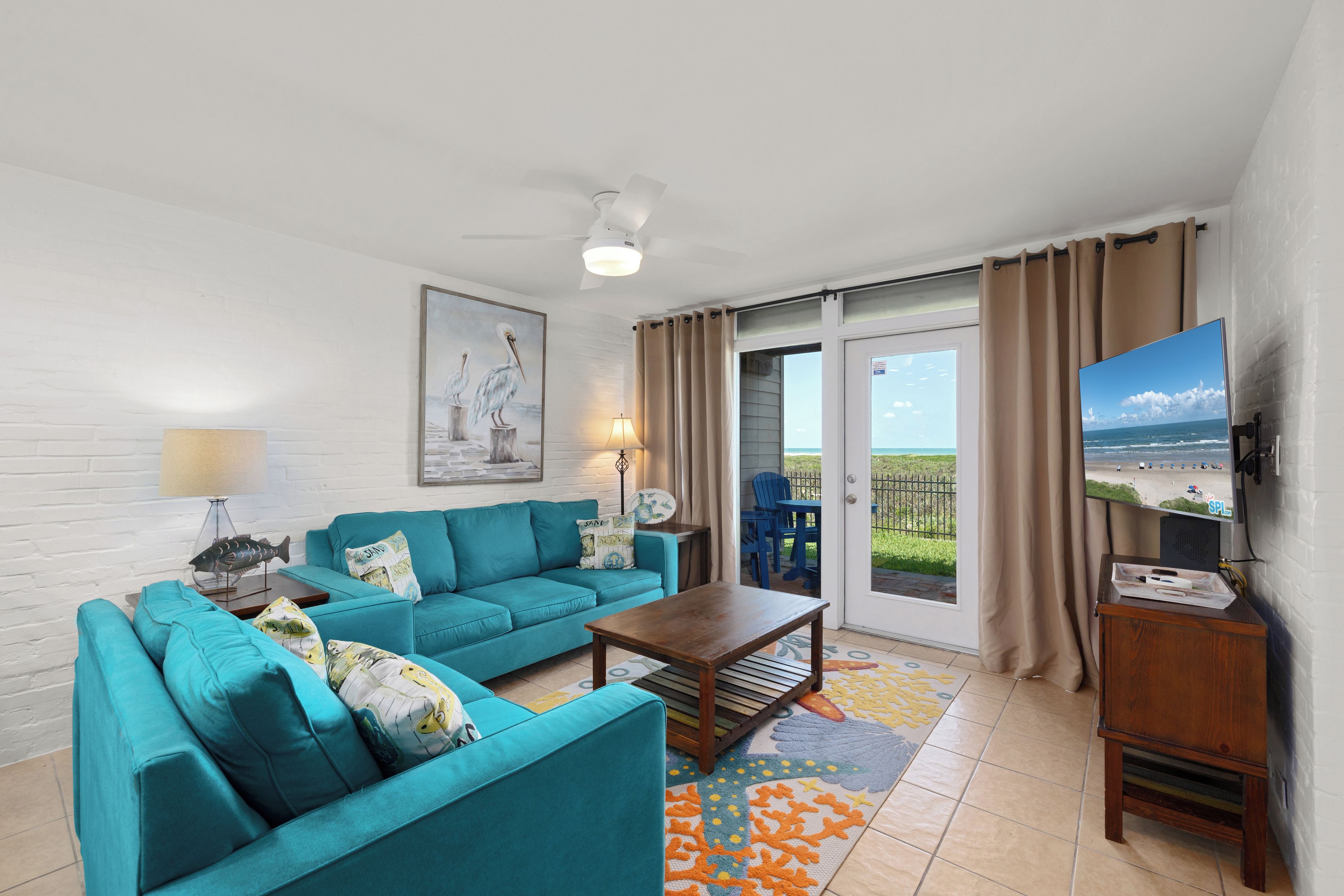A living room with a teal sofa set, a wooden coffee table, a painting of pelicans, a television, and a view of the beach through the sliding glass door.
