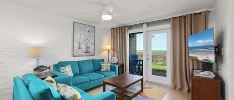 A living room with a teal sofa set, a wooden coffee table, a painting of pelicans, a television, and a view of the beach through the sliding glass door.