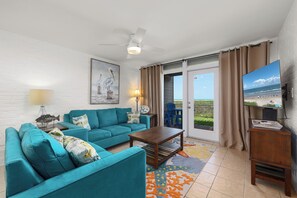 A living room with a teal sofa, a coffee table, a painting of pelicans, a lamp, and a view of the beach through the sliding glass door.