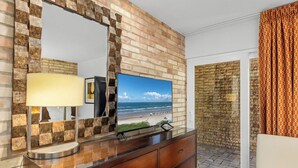 A hotel room with a stone wall, a wooden dresser, a flat-screen TV, and a view of the beach.