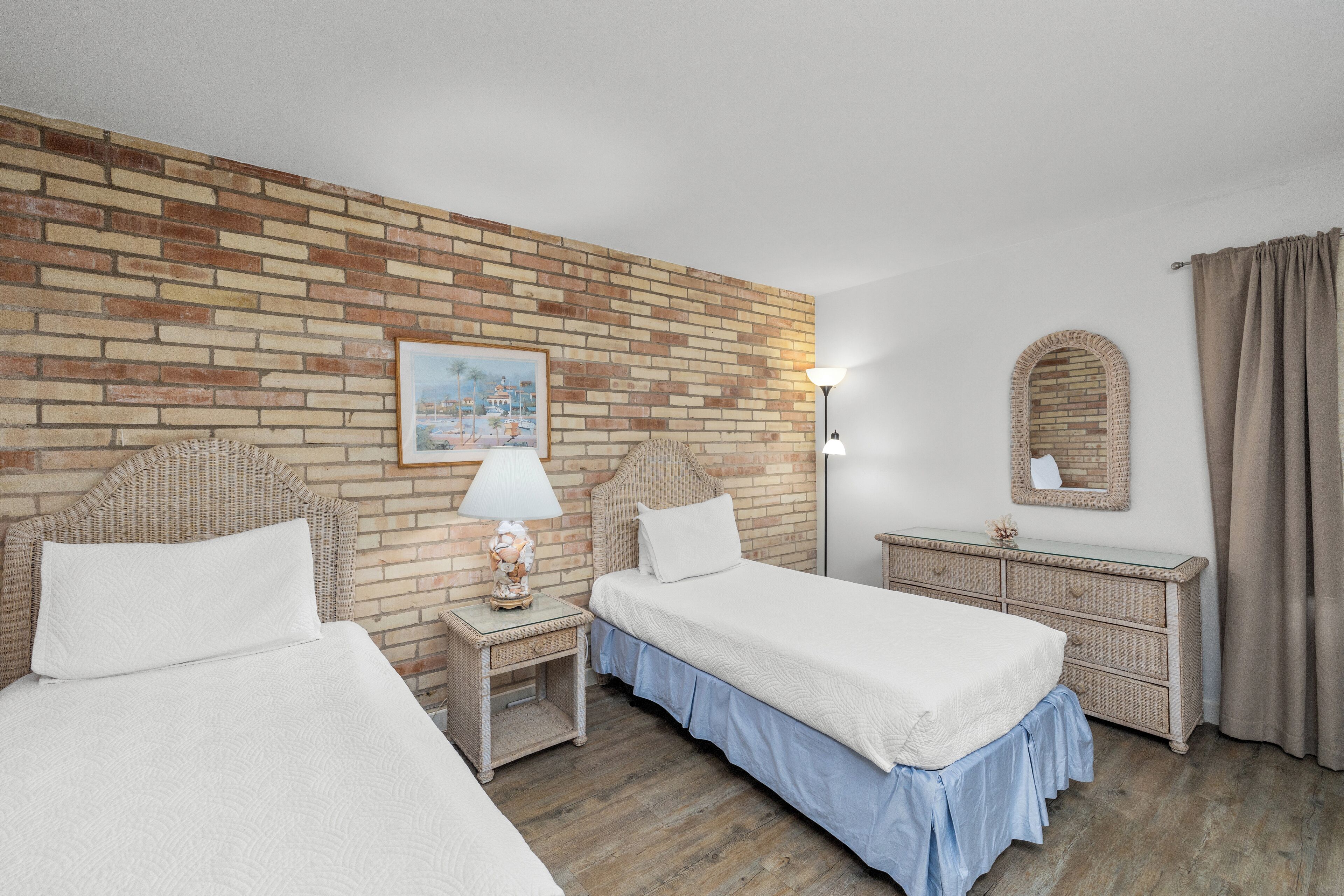A hotel room with two beds, a brick wall, a painting, a lamp, a dresser, and a mirror.