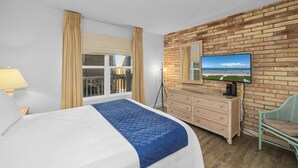 A hotel room with a large bed, a dresser, a chair, a TV on a brick wall, and a window with curtains. - Charming 2-Bedroom Condo. Unbeatable Beach Access! (South Padre Island)