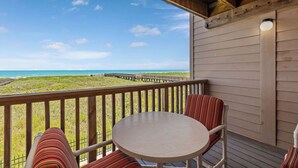 A wooden deck with a round table and striped cushions, overlooking a beach with a wooden pier. - Soothing Seaside Comfort in a 2-Bedroom Escape (South Padre Island)
