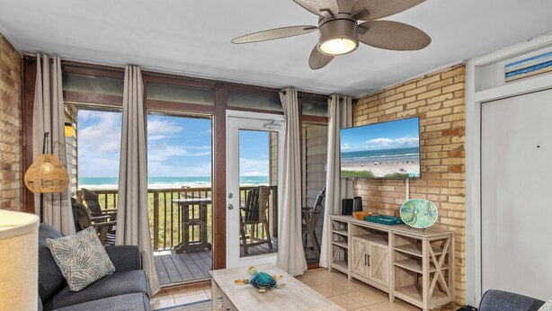 A cozy living area with a sofa, a table, and a TV on a brick wall. There's a balcony with a view of the ocean.
