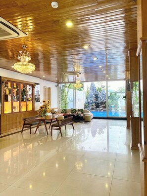 Un hall d’entrée spacieux avec un plafond en bois, un lustre, un coin salon avec une table et des chaises, et une vue sur un élément décoratif aquatique extérieur.