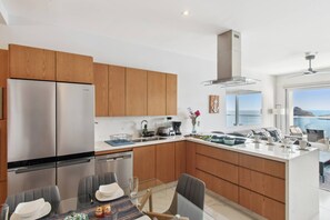 Una cucina moderna con mobili in legno, elettrodomestici in acciaio inossidabile, un'area pranzo con tavolo in vetro e vista sul mare.