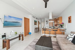 En moderne stue med sofa, sofabord, fjernsyn og et køkkenområde med barstole.