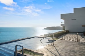 An infinity pool with a view of the sea, a wooden deck, and a modern building with a flat roof. - Beautiful Long stay Unit with Premium Ameneties (Mazatlan)