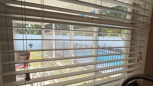 A kitchen with a window with horizontal blinds, a sink, and a view of a backyard with a pool.