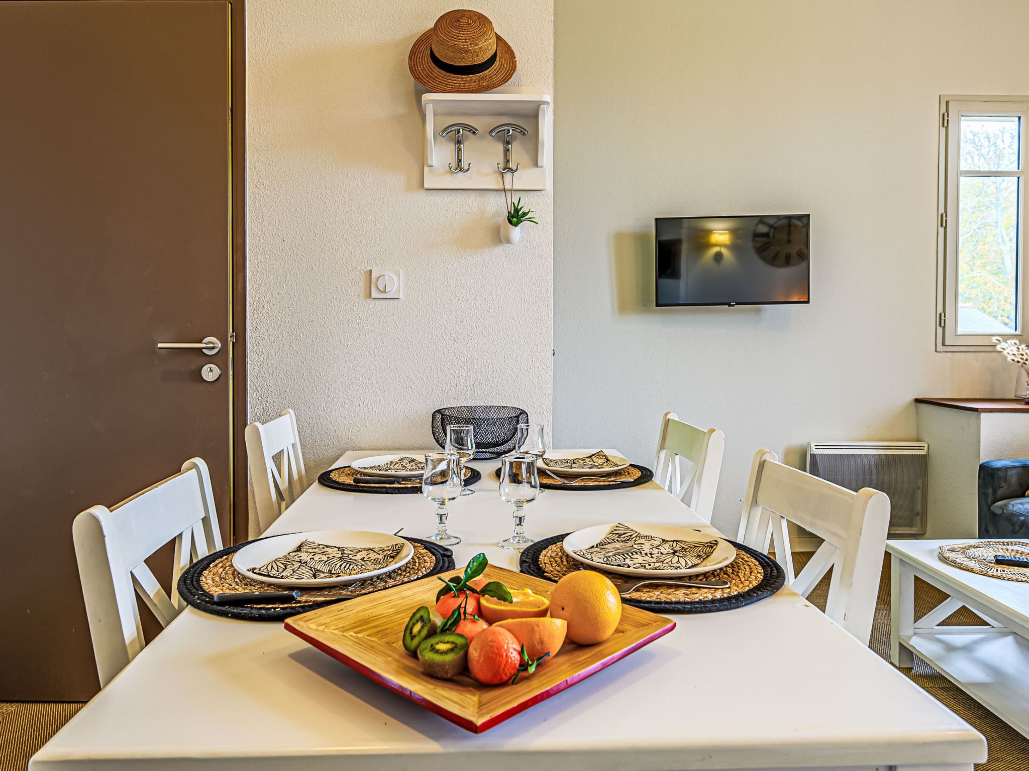 Un comedor con una mesa preparada para cuatro personas, un televisor montado en la pared y una puerta a la izquierda.