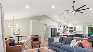 Ein geräumiges Wohnzimmer mit grauem Sofa, braunem Sessel und Couchtisch. Im Hintergrund ist eine Küche mit weißen Schränken sowie ein Essbereich mit Tisch und Stühlen zu sehen.