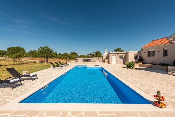 Une piscine rectangulaire avec des chaises longues et un panier de fruits sur le côté droit.