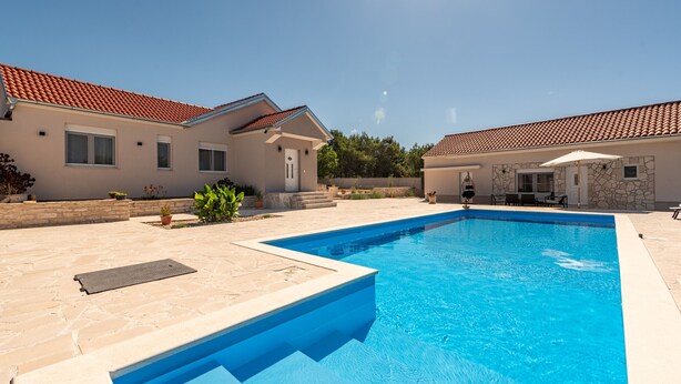 Une piscine à l’eau d’un bleu limpide, entourée d’une terrasse en pierre, et jouxtant un bâtiment dont le toit est recouvert de tuiles rouges.