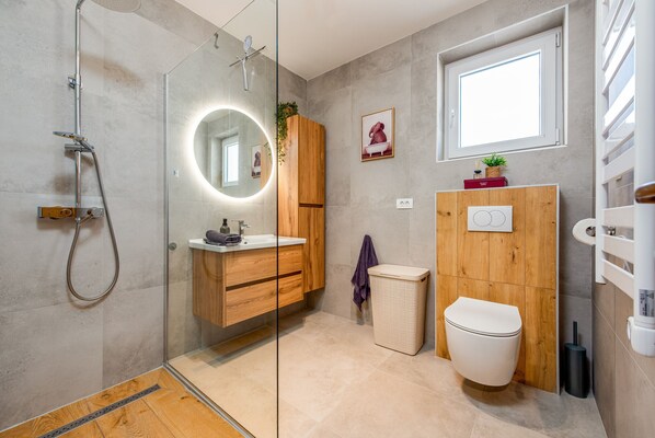 A modern bathroom with a glass shower enclosure, a wooden vanity, a toilet, and a window.
