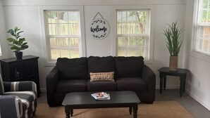 A living room with a dark sofa, a coffee table, and two side tables. There is a 'Welcome' sign on the wall and a plant on one of the side tables.