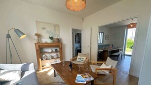 A cozy living room with a sofa, a wooden coffee table, and a chair. There is a bookshelf with decorative items and a painting on the wall.