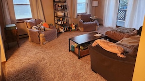 A cozy living room with a sofa, armchair, and a coffee table. There is a bookshelf filled with books and decorative items.