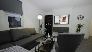 Una sala de estar moderna con un sofá gris, una mesa de centro de cristal, un sillón negro, un televisor montado en la pared y un reloj.