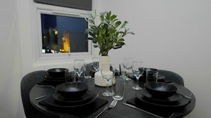 Una mesa de comedor dispuesta con platos, fuentes y cubiertos negros, un florero blanco con plantas verdes, y una ventana con vista a la ciudad.