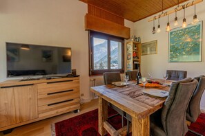 Un salon moderne avec une table à manger en bois, des chaises, un téléviseur à écran plat et une grande fenêtre donnant sur une vue imprenable sur les montagnes.