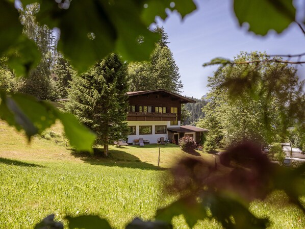 Un chalet de dos pisos con techo inclinado, rodeado de un paisaje verde exuberante y árboles.