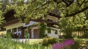 A two-story chalet with a balcony, surrounded by lush greenery and trees.
