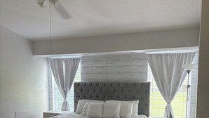 A bedroom with a large bed, a nightstand, a ceiling fan, and a window with curtains.