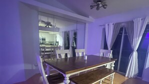 A dining area with a wooden table and white chairs, a ceiling fan, and a view of a kitchen and balcony through a large window.