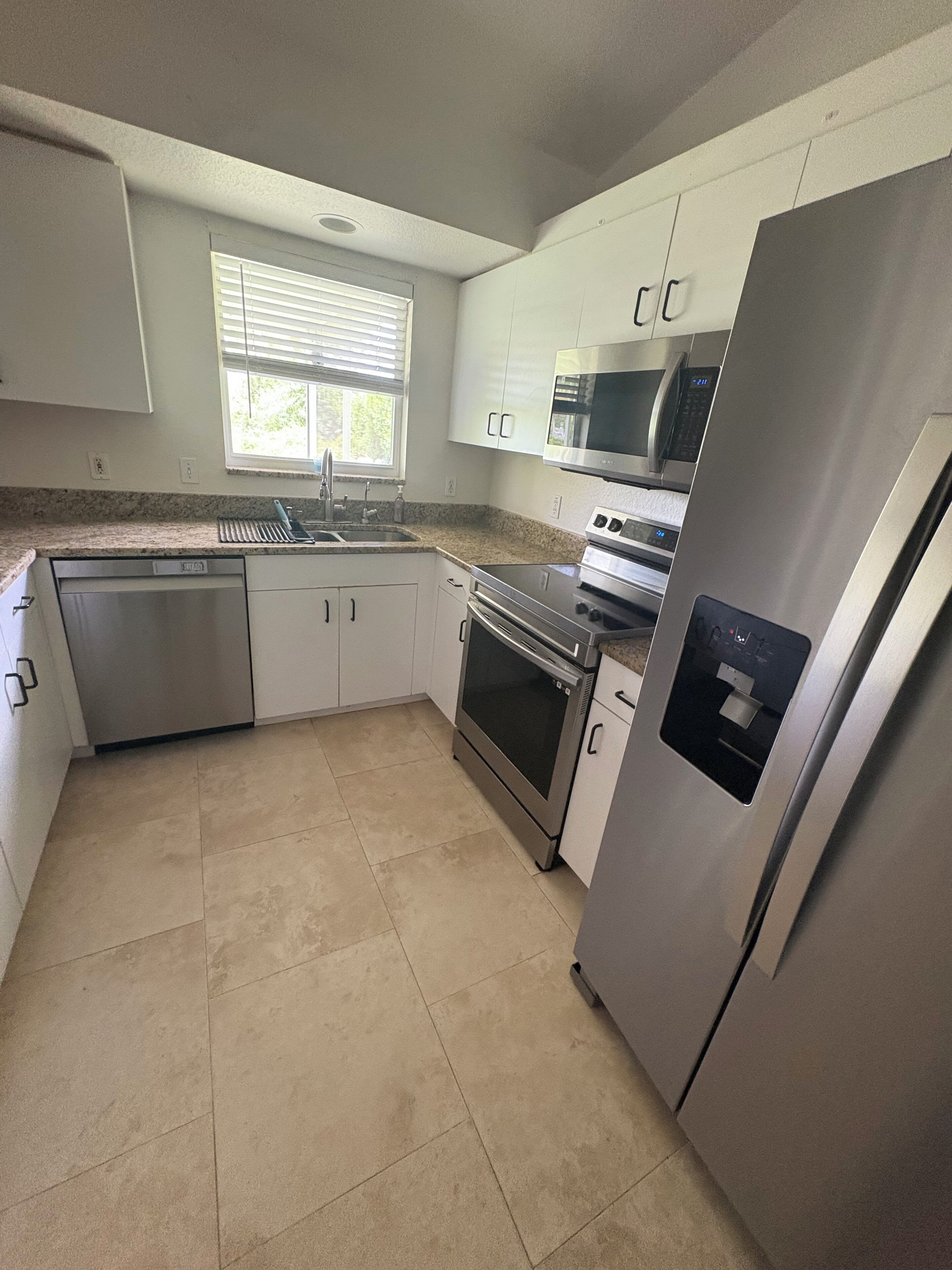 A modern kitchen with white cabinets, stainless steel appliances, and a window with blinds.