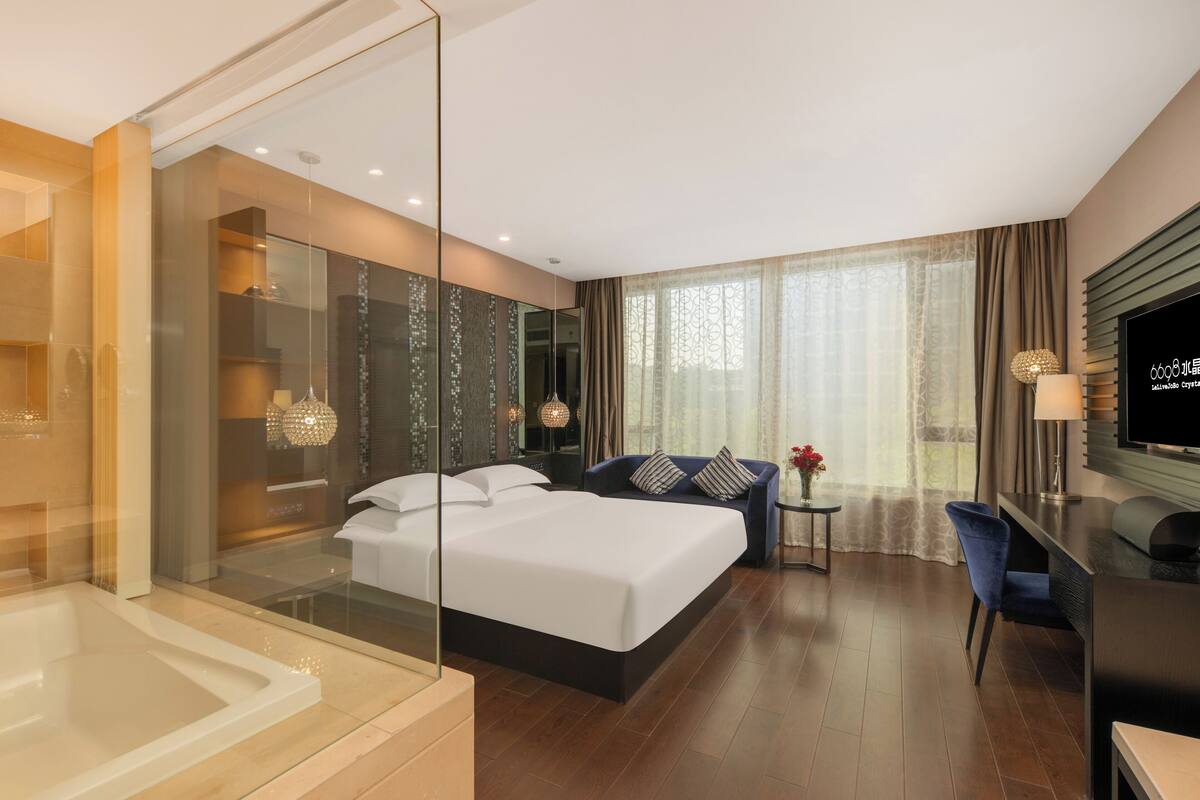 A modern hotel room with a large bed, a glass-enclosed shower area, a seating area with a sofa and armchairs, and a television on the wall.