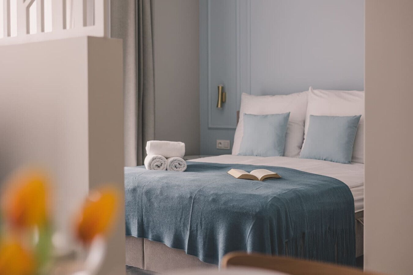 Quarto com cama, colcha azul, duas almofadas, toalha enrolada e um livro aberto.