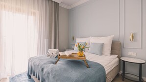 Ein Hotelzimmer mit einem Bett, einem Nachttisch, einem kleinen runden Tisch, einem Fenster mit Vorhängen sowie einem Tablett mit Handtüchern und einer Blumenvase.