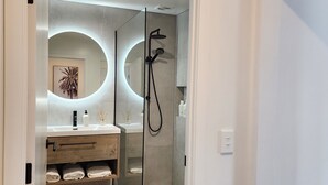 A modern bathroom with a glass-enclosed shower, a vanity with a mirror, and a towel rack. - Rooftop Apartments (Raglan)