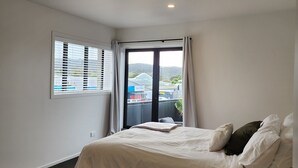 A bedroom with a large bed, a window with blinds, a ceiling light, and a view of buildings outside. - Rooftop Apartments (Raglan)