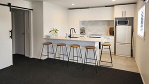 A modern kitchen with white cabinets, a black faucet, a microwave, and a refrigerator. There are four stools and a potted plant on the counter. - Rooftop Apartments (Raglan)