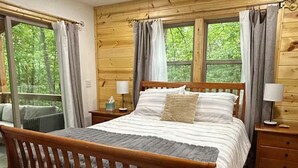 A wooden cabin bedroom with a bed, nightstands, a ceiling fan, and large windows with curtains.