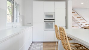 A modern kitchen with white cabinetry, a built-in oven, and a dining area with a high wooden table and woven chairs.