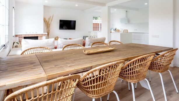 A spacious dining area with a long wooden table and wicker chairs, a television mounted on the wall, and a kitchen in the background.
