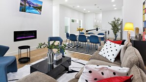 A modern living room with a flat-screen TV, a coffee table with books and a vase, a sofa with patterned cushions, and a dining area with blue chairs and a black and white rug. - Spacious 4-bedroom house Game Room & Spa in fantastic Dallas 11 (Dallas)