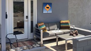 Un patio cubierto con puerta corrediza de vidrio, un sofá con cojines a rayas, una mesita y un cartel de advertencia de vaca en la pared.