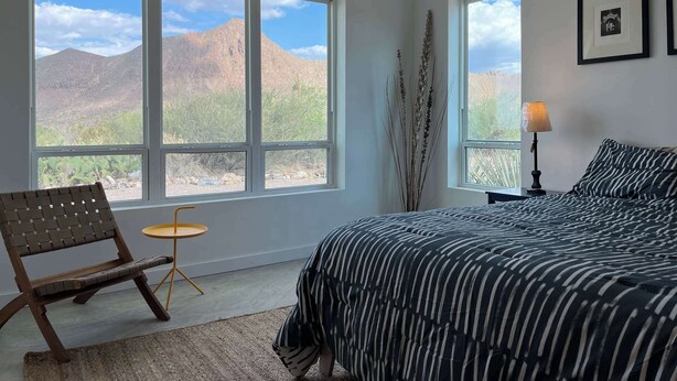 Un dormitorio con una cama, una silla, una mesita y una lámpara. Cuenta con ventanas amplias y vistas a la montaña.