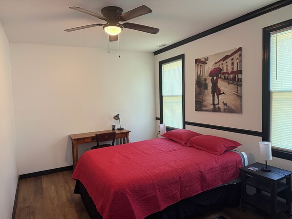 Un dormitorio con cama, escritorio, silla, ventilador de techo y dos ventanas con persianas.