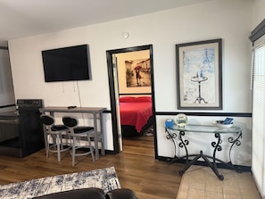 Ein kompakter Wohnbereich mit Küchenzeile, Essbereich, Wandfernseher und Bett mit roter Decke.