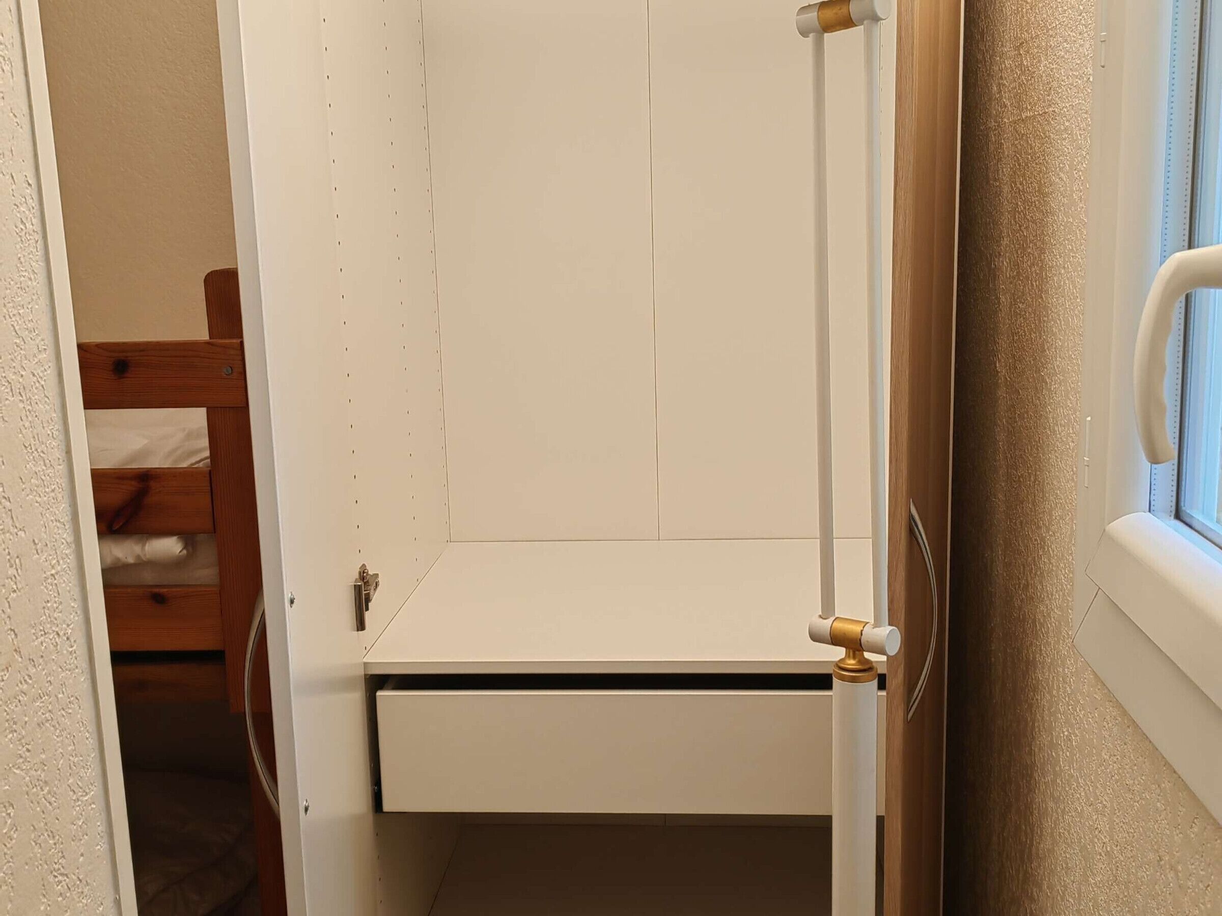 Un armario blanco con puerta corredera y un cajón pequeño, situado junto a una ventana con marco blanco.