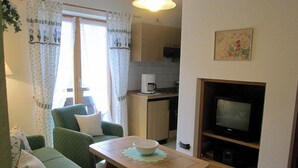 Un espacio habitable compacto con un sofá verde, una pequeña mesa de comedor con un bol, una zona de cocina con microondas y un televisor contra la pared.