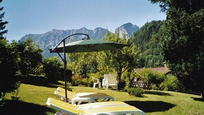Un jardín con tumbonas y una mesa bajo una pérgola, rodeado de árboles y montañas al fondo.