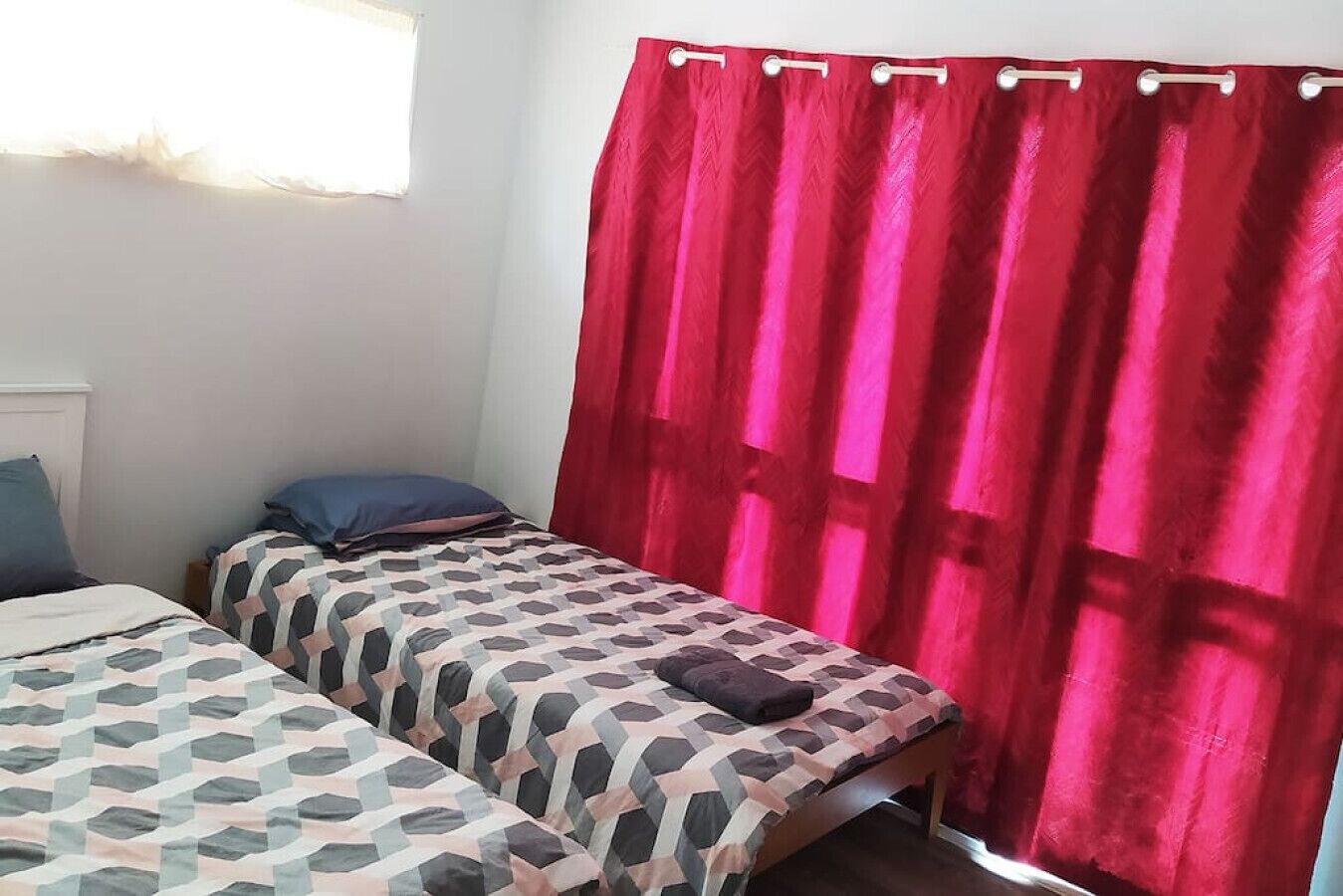Une chambre avec deux lits, chacun agrémenté de draps à motifs, d’un rideau rouge et d’une fenêtre laissant entrer la lumière naturelle.