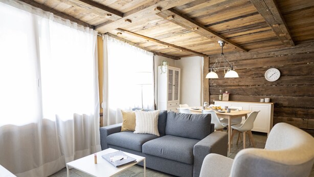 A modern living room with a grey sofa, a small white table, and a dining area with a wooden table and chairs. - Chasa Rustica – Modern 3-Room Apartment with Traditional Charm in Sent (Sent)