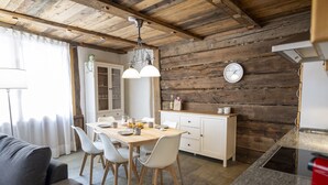 A cozy kitchen with wooden beams, a dining table with chairs, a clock on the wall, and a cabinet with dishes. - Chasa Rustica – Modern 3-Room Apartment with Traditional Charm in Sent (Sent)